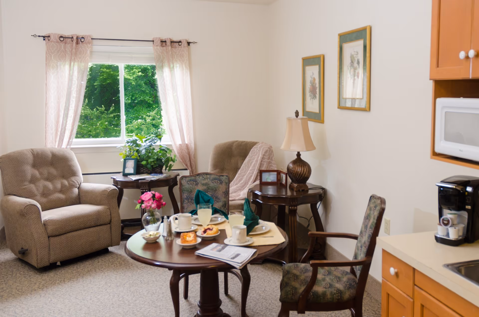 A cozy living room area with two beige armchairs, a round wooden table set with plates, cups, and glasses, and two wooden side tables. One side table has a lamp and framed photos, while the other has a plant and a framed photo. There is a window with pink patterned curtains showing green trees outside. A coffee maker and microwave are visible on the right on a countertop with wooden cabinets below.