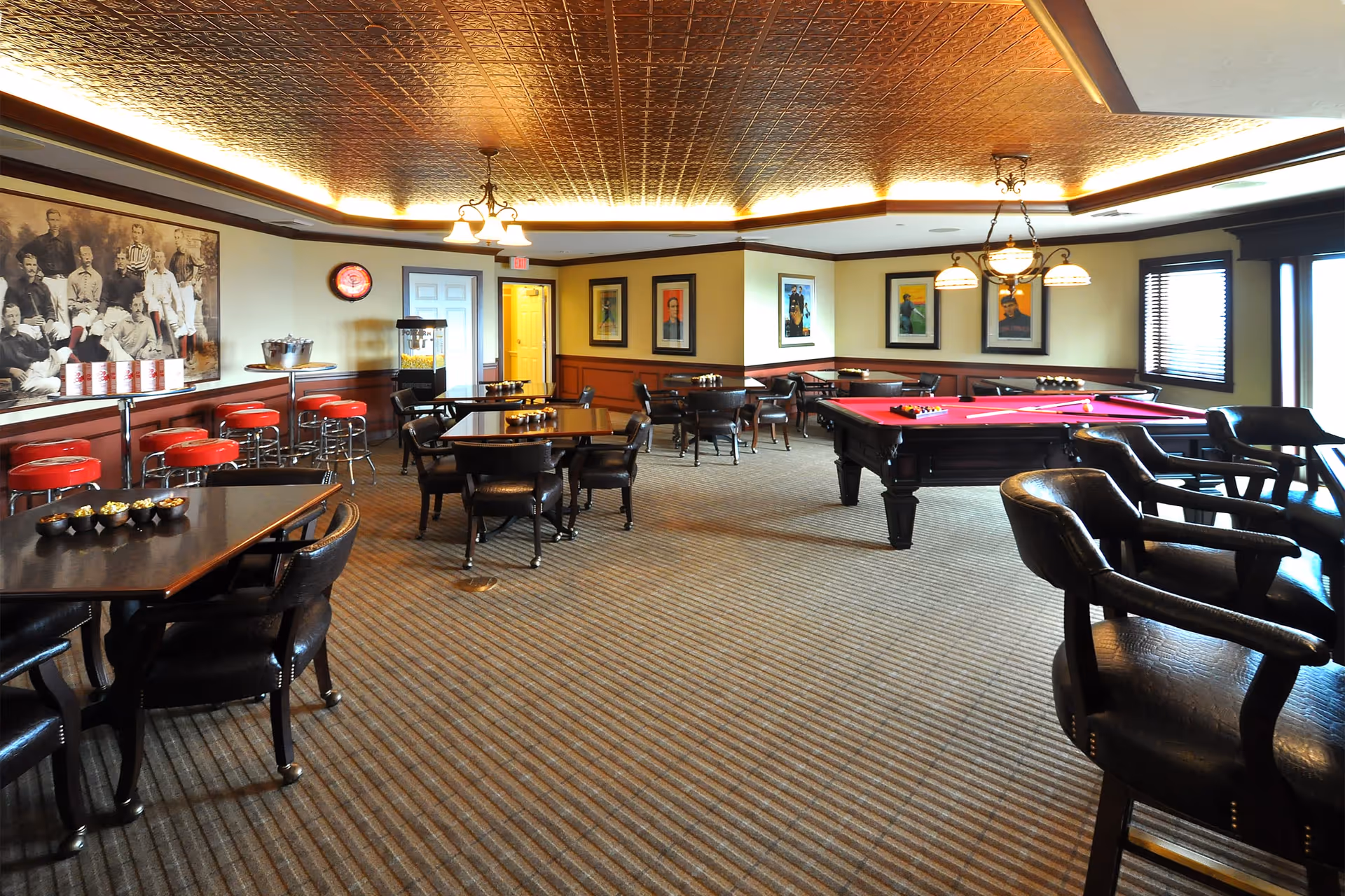 A spacious recreational room with multiple tables and chairs, a red pool table with balls set up, a popcorn machine, and red bar stools along a counter. The room has a patterned carpet, framed artwork on the walls, and a decorative ceiling with warm lighting.