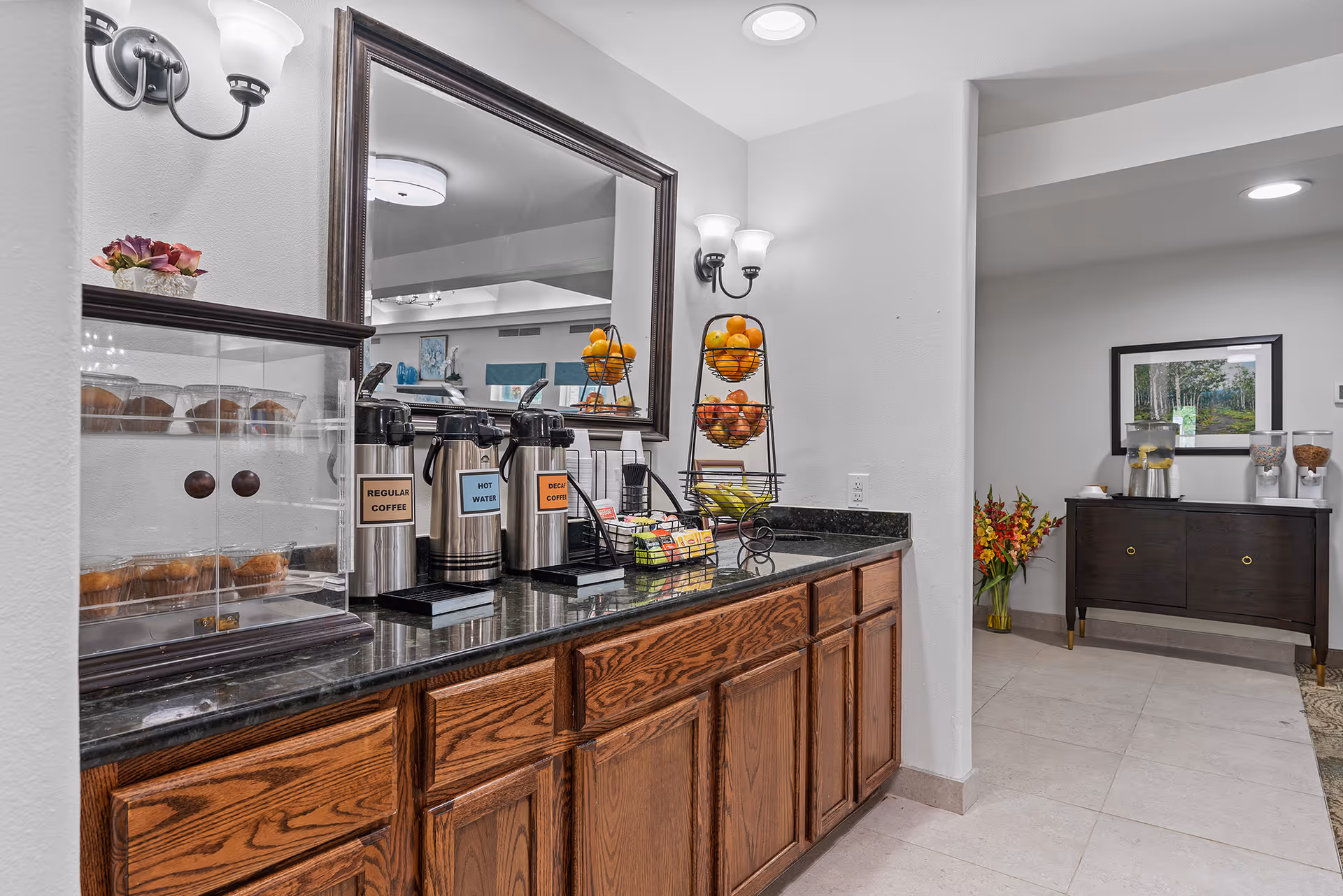A coffee and snack station in a senior living facility with three beverage dispensers labeled Regular Coffee, Hot Water, and Decaf Coffee, a display case with muffins, a tiered fruit basket with oranges and bananas, and a countertop with various coffee supplies. The area features wooden cabinets, a large mirror, wall-mounted light fixtures, and a view into an adjacent room with a dark wooden sideboard, cereal dispensers, a water dispenser, a framed nature picture, and a vase with flowers.