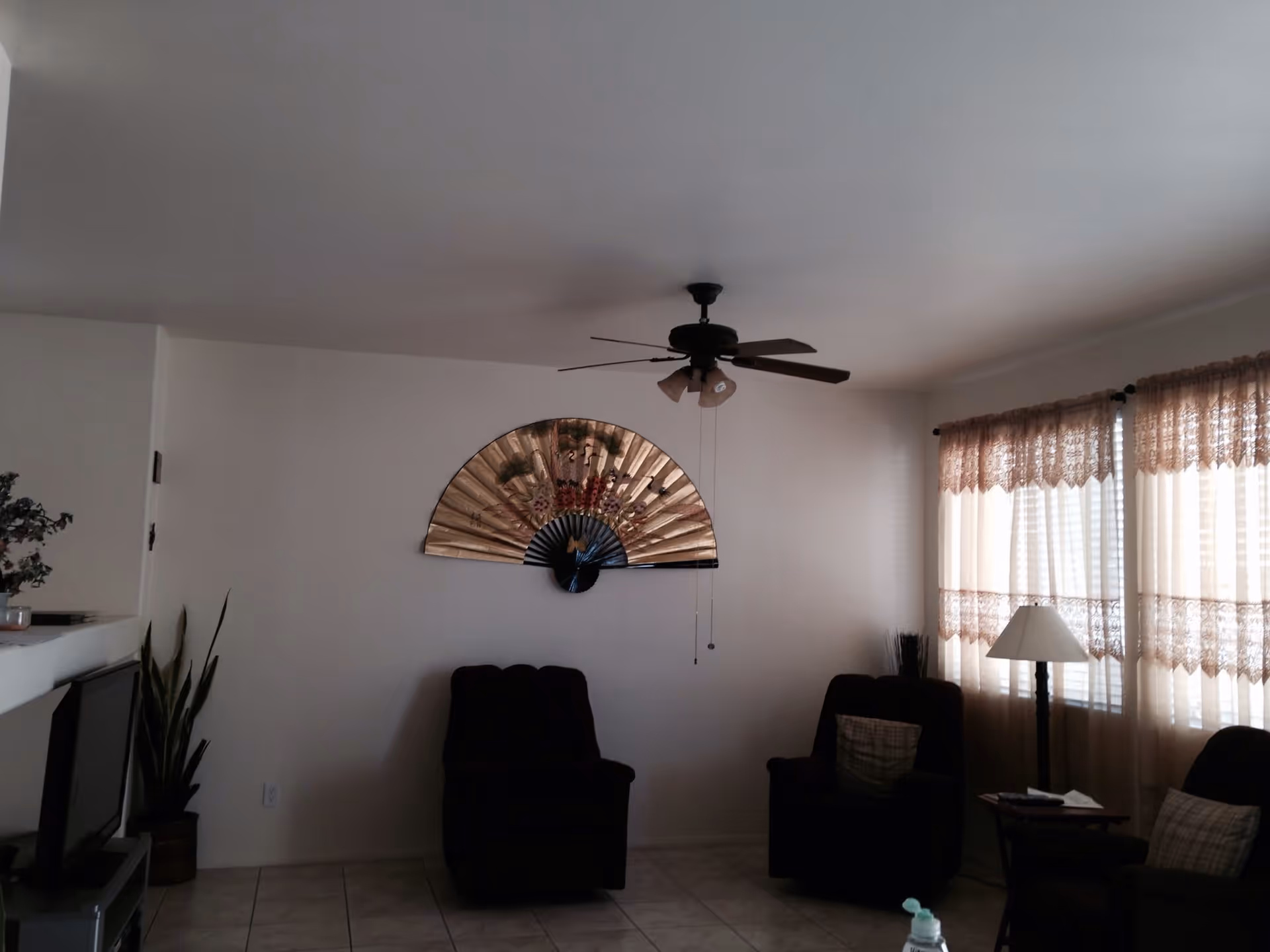 A living room with two dark armchairs, a floor lamp, a small side table, and a large decorative fan mounted on a white wall. The room has tiled flooring, a ceiling fan with lights, and windows covered with lace curtains allowing natural light to enter.