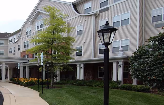Front exterior of a multi-story senior living building with a covered entrance, lampposts, walkway, and landscaped lawn.