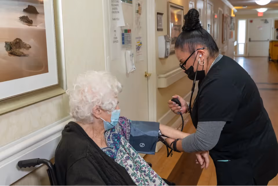 A healthcare worker wearing a black mask and scrubs is taking the blood pressure of an elderly woman with white hair who is seated in a wheelchair in a hallway of a care facility.