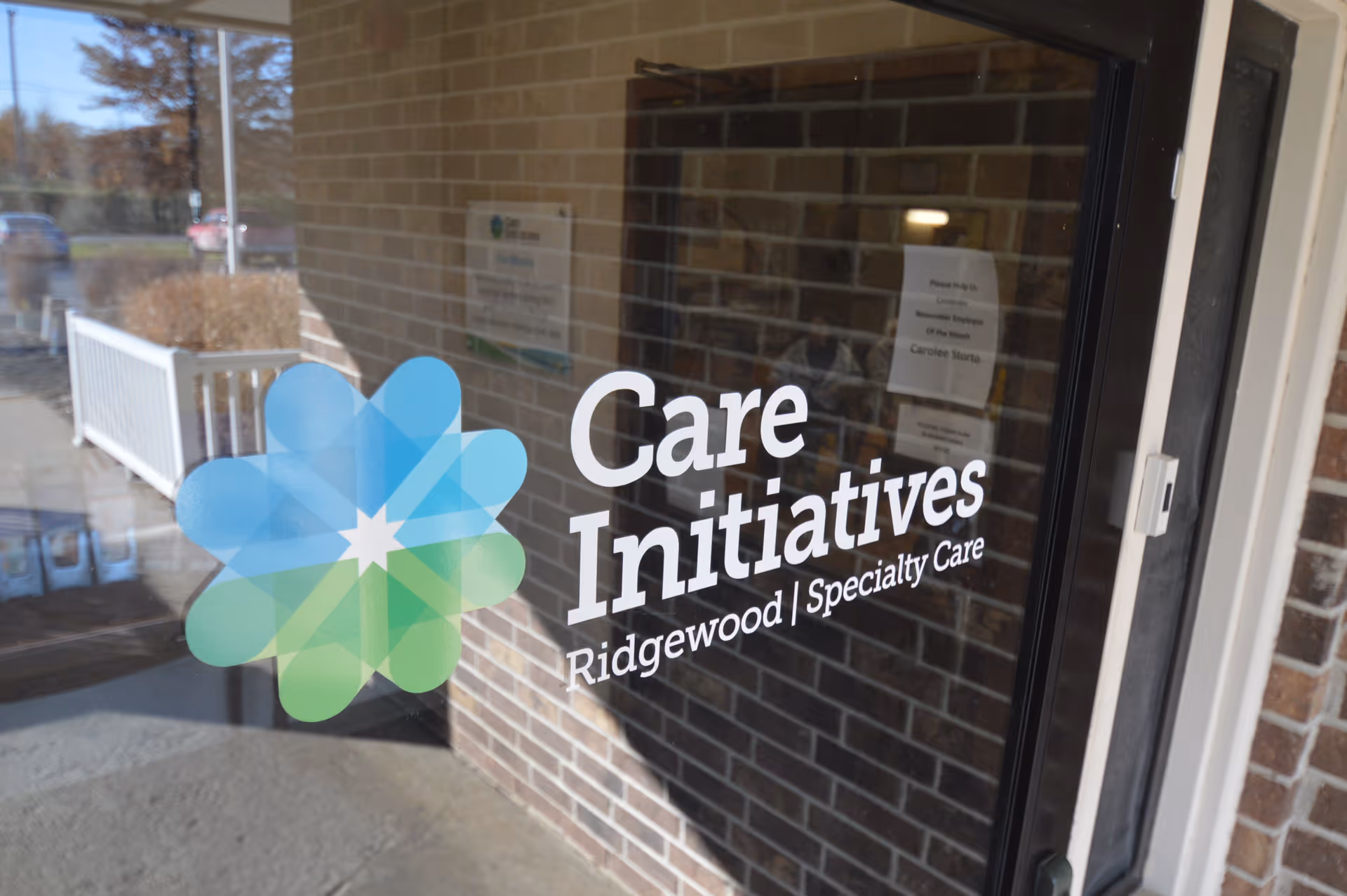 Glass door with the logo and text 'Care Initiatives Ridgewood | Specialty Care' on it, reflecting the exterior brick wall and a white railing outside.