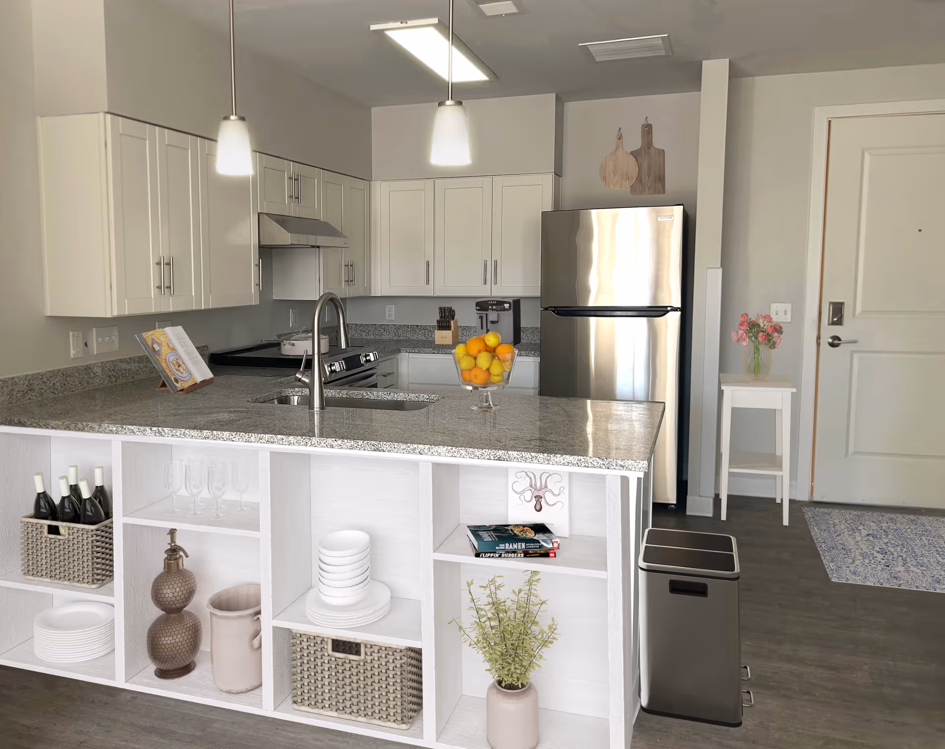 Modern kitchen with white cabinets, stainless steel refrigerator and stove, granite countertops, and an island with open shelving containing dishes, wine bottles, and decorative items. Two pendant lights hang above the island, and a small table with a vase of flowers is near the entrance door.