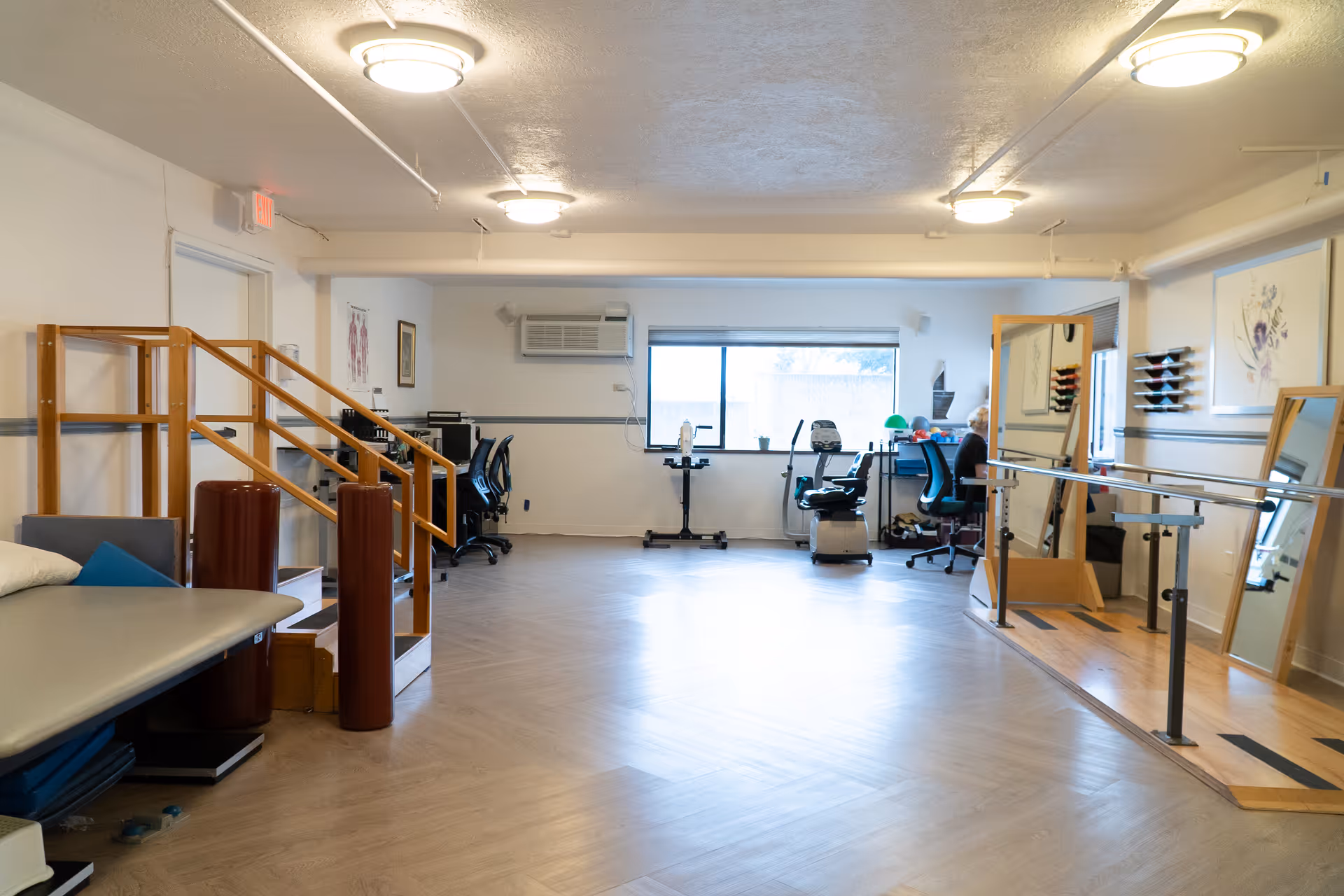 A spacious rehabilitation or physical therapy room with wooden handrails, exercise equipment including stationary bikes, large mirrors, and a padded therapy table. The room has bright overhead lighting and a large window letting in natural light.