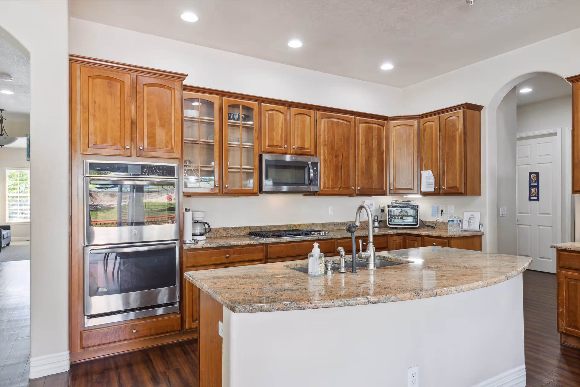 A modern kitchen with wooden cabinets, granite countertops, and stainless steel appliances including a built-in oven and microwave. The kitchen features a large island with a sink and faucet, and recessed lighting in the ceiling. There is an arched doorway leading to another room and a glimpse of a living area with windows in the background.