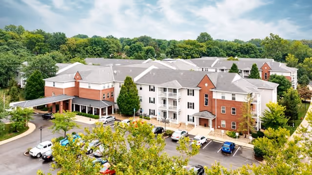 Aerial view of The Emerson at St. Peters senior living facility showing a large multi-story building with a combination of red brick and white siding exterior, surrounded by trees and a parking lot with several cars. The sky is partly cloudy.