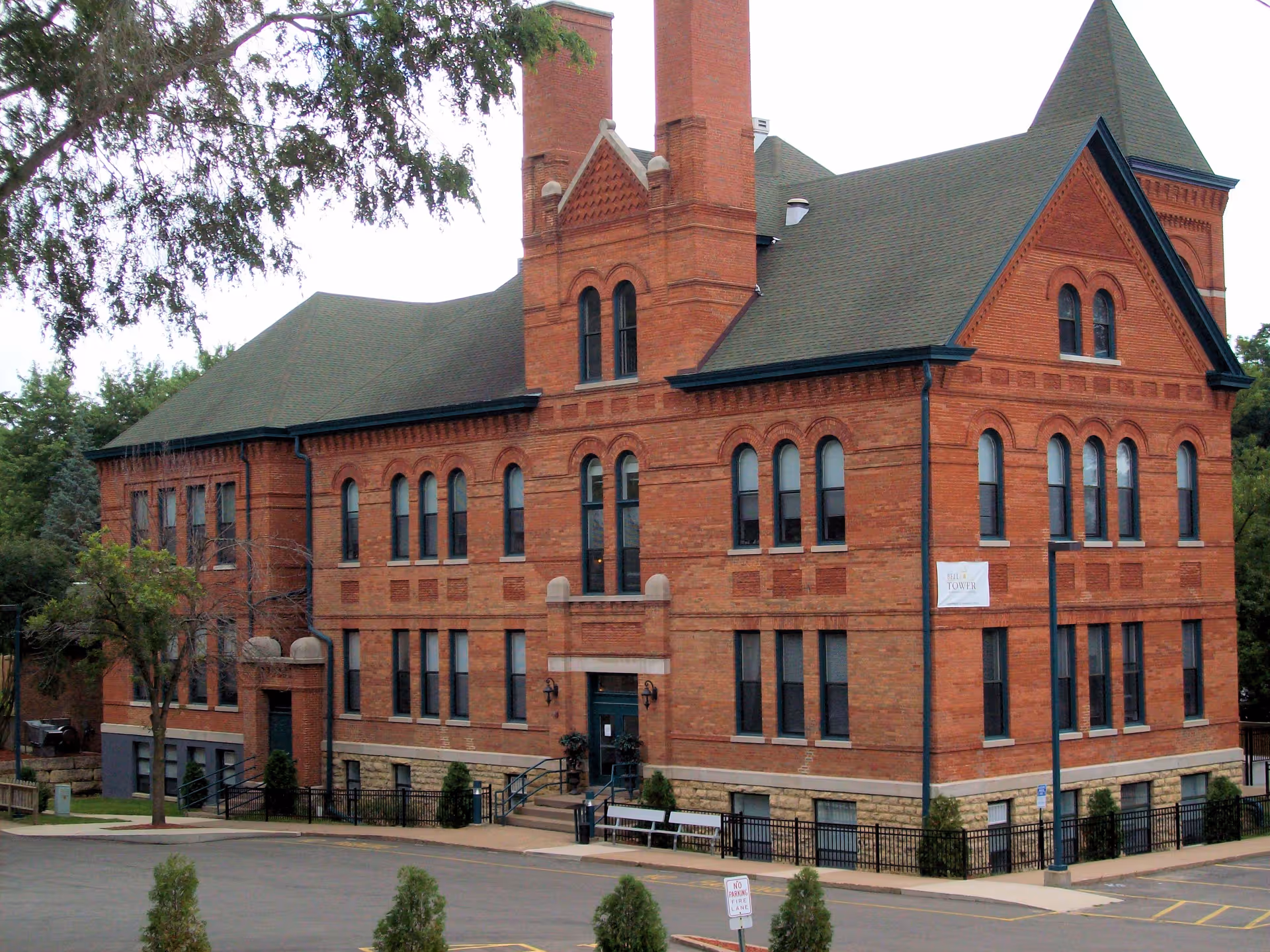 A large, historic red brick building with multiple arched windows and a steep green roof, surrounded by trees and a parking lot in front.