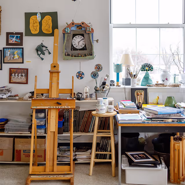 Sunlit interior art studio with an easel, stools, shelves of books and supplies, and artwork on the wall beneath a large window.