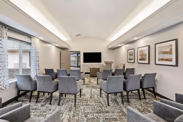 Communal seating room with rows of gray upholstered chairs facing a wall-mounted TV and podium.