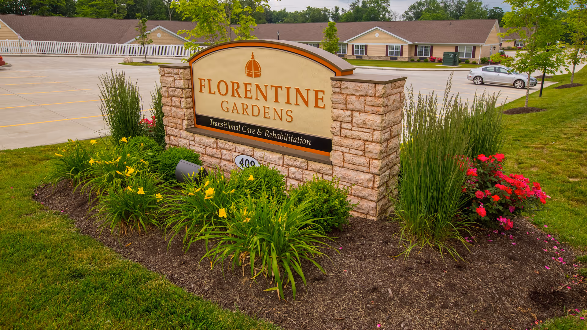 Stone sign for Florentine Gardens Transitional Care & Rehabilitation surrounded by landscaped flowers and greenery, with a parking lot and single-story buildings in the background.
