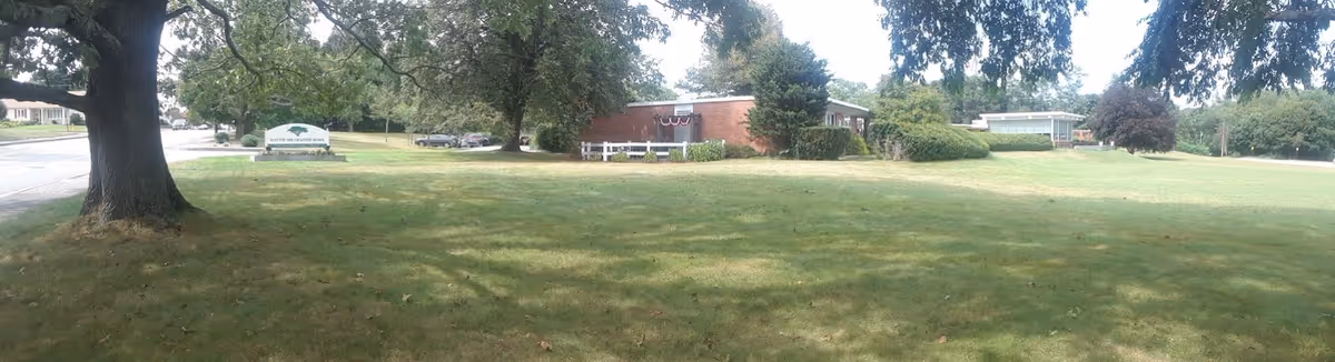 A wide grassy lawn with scattered trees and bushes in front of a low brick building. There is a sign on the lawn that reads 'Hattie Ide Chaffee Home'. The sky is partly cloudy and the area appears to be a quiet suburban neighborhood.