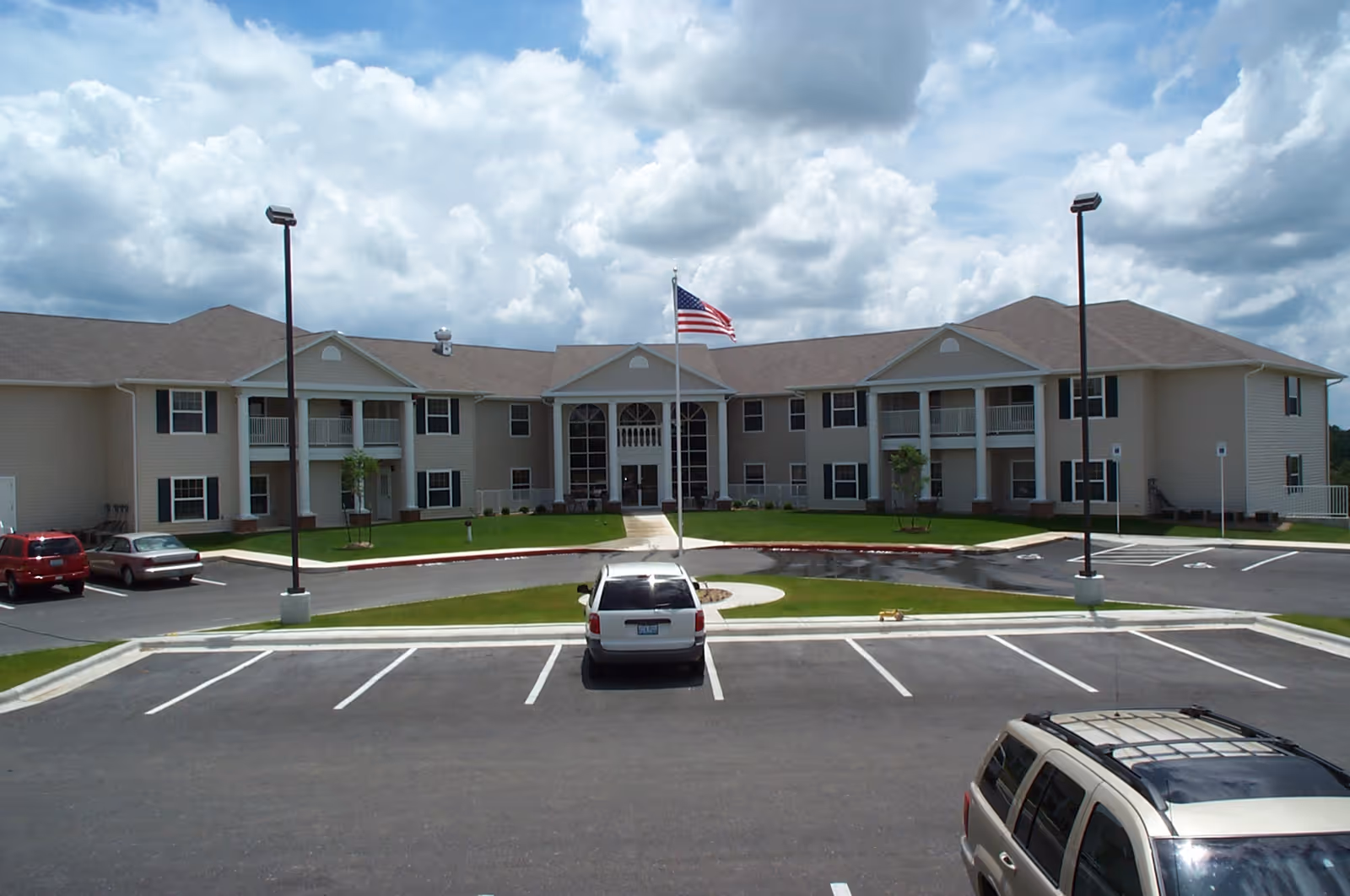 Exterior view of a two-story beige retirement community building with a central entrance, balconies, and an American flag on a flagpole in front. Several cars are parked in the parking lot under a partly cloudy sky.
