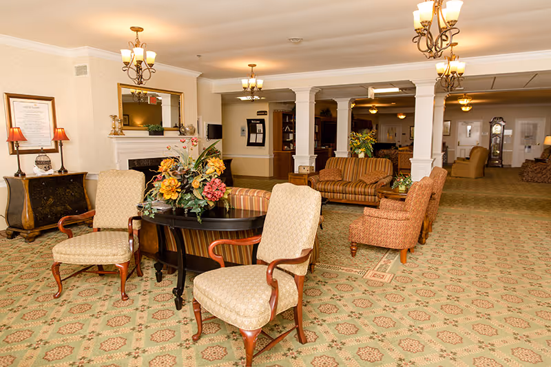A spacious and warmly lit senior living facility common area with patterned carpet, multiple upholstered chairs and sofas arranged around small tables, decorative floral centerpieces, chandeliers hanging from the ceiling, and a fireplace with a large mirror above it.
