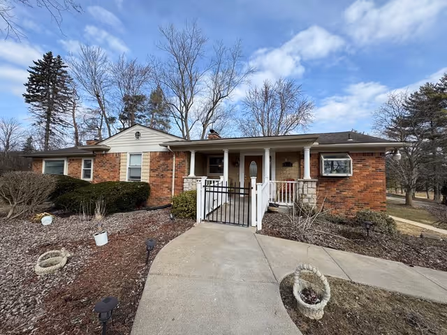 Single-story brick building with a covered front porch and a gated entrance. The building is surrounded by leafless trees and some shrubs, with a concrete pathway leading to the entrance. The sky is partly cloudy.
