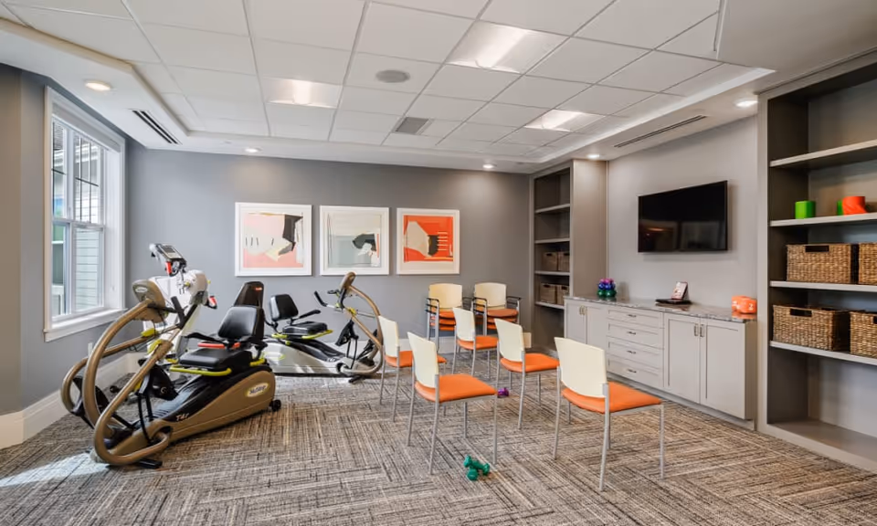 Well-lit interior activity room with recumbent exercise bikes, several chairs arranged in rows, wall art, shelving and a TV.