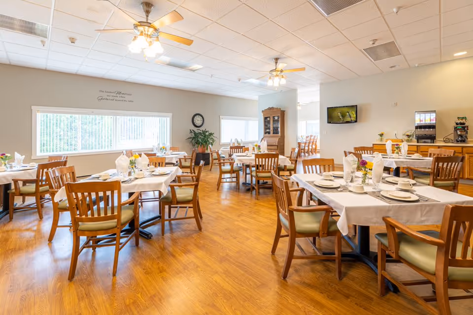 A bright and spacious dining room with wooden floors, multiple tables set with white tablecloths, plates, cups, and napkins. Wooden chairs with green cushions surround the tables. The room has large windows with vertical blinds, ceiling fans with lights, a wall clock, a plant, a wooden cabinet, a wall-mounted TV, and a beverage station with a coffee machine and juice dispenser.