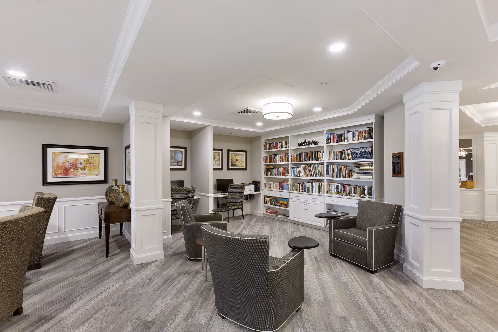 A cozy and well-lit common area in a senior living facility featuring a built-in white bookshelf filled with books, a welcome sign, and a few comfortable armchairs with small attached tables arranged around the room. The space has light gray wood flooring, white paneled columns, and framed artwork on the walls. There are also two computer workstations with chairs in a small alcove.