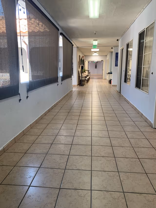 A long, clean hallway with tiled floor and white walls. On the left side, there are windows covered with dark roller shades, and on the right side, there are several windows and doors. At the far end of the hallway, there are chairs and some plants near the wall. The ceiling has fluorescent lights and an exit sign is visible in the distance.