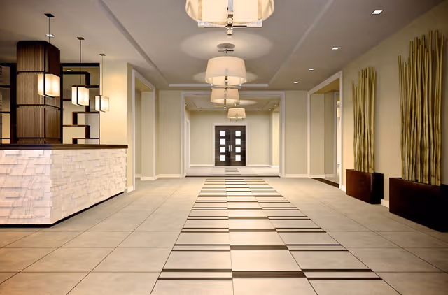 A spacious, modern interior hallway with tiled flooring featuring a striped pattern. The hallway is well-lit with multiple ceiling lights and pendant lamps. On the left side, there is a reception desk with a textured front panel and decorative shelving behind it. On the right side, tall decorative bamboo plants are placed in rectangular planters against the wall. At the end of the hallway, double doors with glass panels are visible.