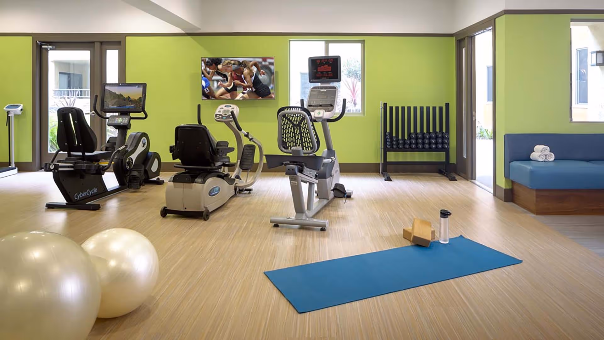 A brightly lit exercise room with green walls featuring two recumbent exercise bikes, a rack of dumbbells, two large exercise balls, a blue yoga mat with a water bottle and yoga blocks, and a blue cushioned bench with rolled towels. A TV mounted on the wall shows an image of runners at the starting line.