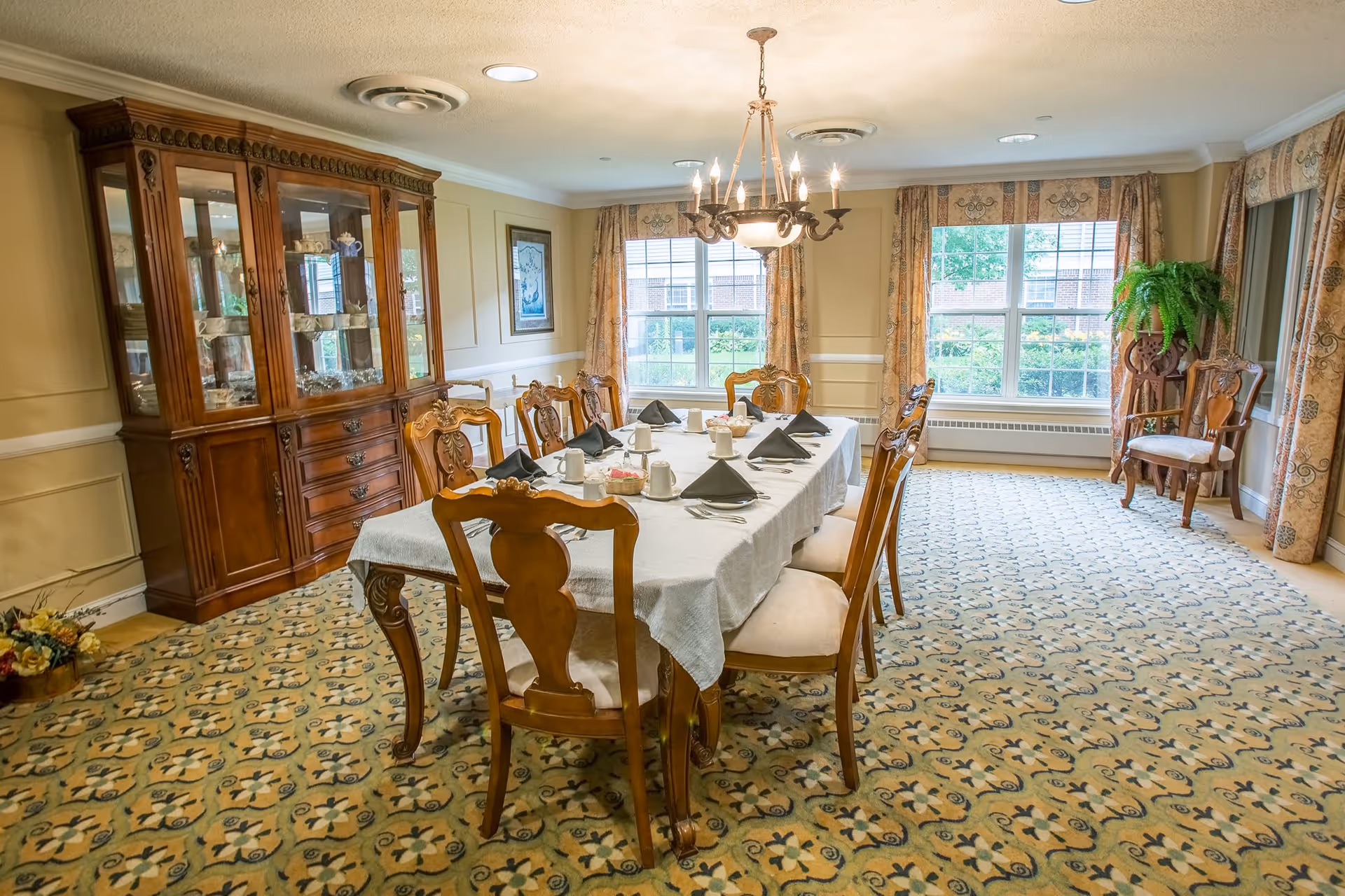 A formal dining room with a long wooden table covered with a white tablecloth, set with cups, plates, silverware, and black folded napkins. The room features ornate wooden chairs, a large wooden china cabinet filled with dishes, patterned carpet, large windows with floral curtains, and a chandelier hanging from the ceiling.