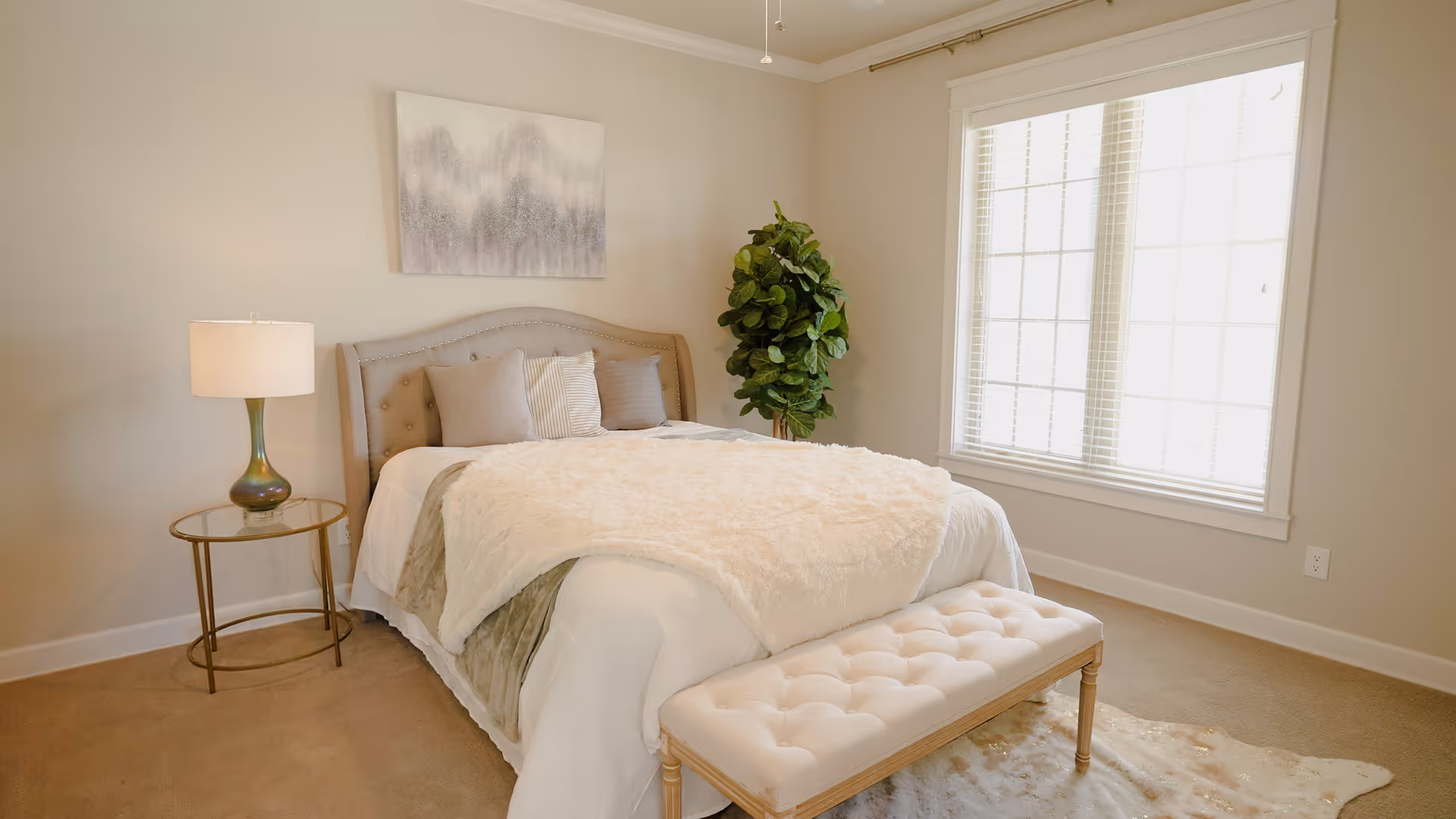 A cozy bedroom with a neatly made bed featuring a tufted headboard, several pillows, and a fluffy white blanket. A small round glass-top side table with a lamp is positioned to the left of the bed. A cushioned bench sits at the foot of the bed. There is a large window with white blinds on the right wall and a green leafy plant in the corner. A soft rug is partially visible on the floor.