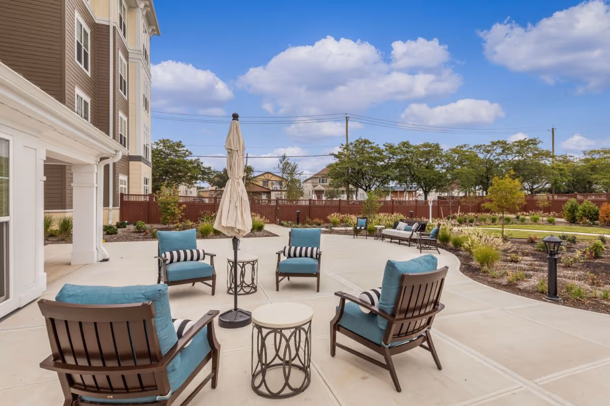 Outdoor patio with blue-cushioned chairs, small tables, a closed umbrella, and landscaped grounds beside a multi-story building.