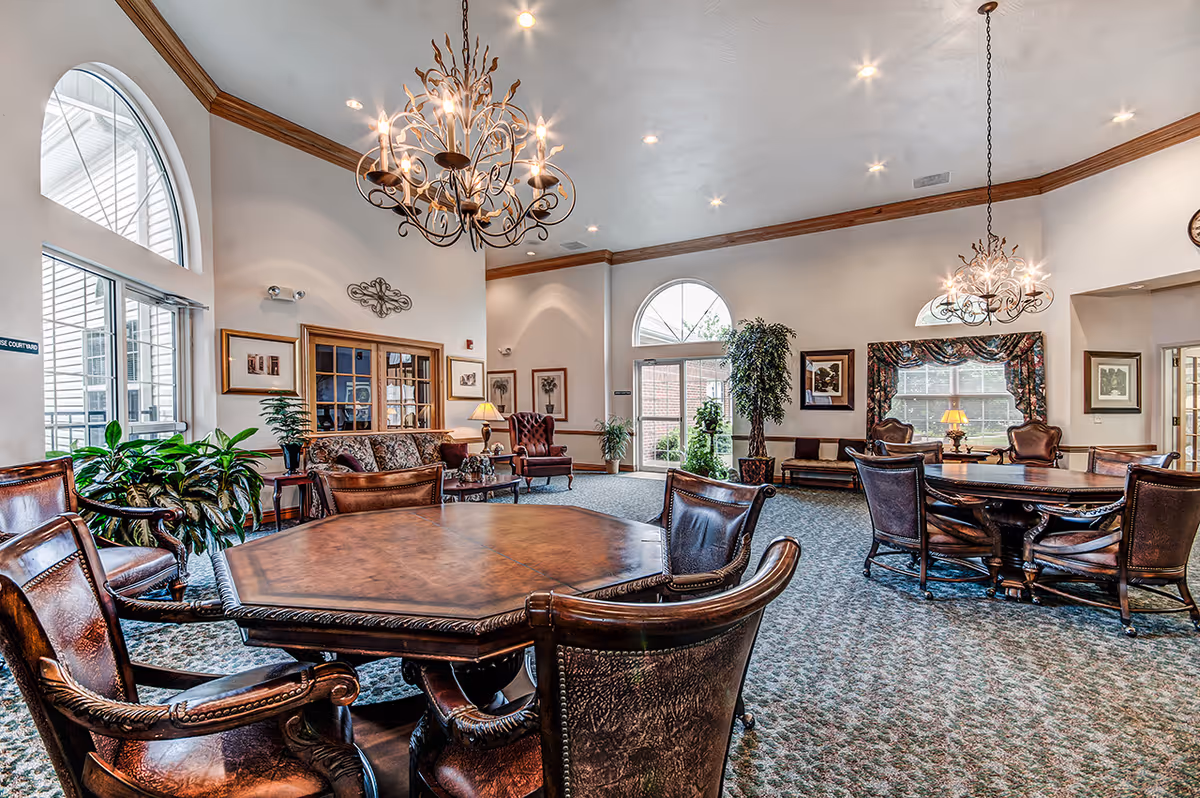 Bright communal lounge with ornate chandeliers, leather chairs around wooden tables, potted plants, and large arched windows.