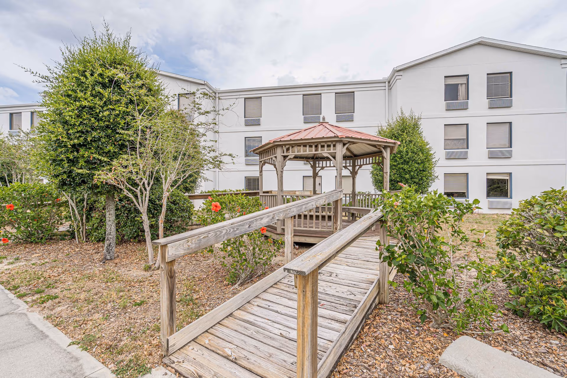 Outdoor garden area with a wooden ramp leading to a gazebo surrounded by bushes and trees, with a multi-story building in the background under a cloudy sky.