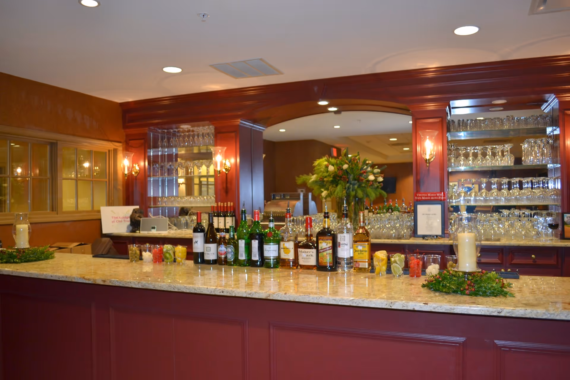 A well-lit bar area with a granite countertop displaying various bottles of liquor and garnishes. Behind the bar, there are shelves filled with glassware and a large mirror reflecting the room. The bar has dark wood paneling and warm lighting fixtures on the walls.