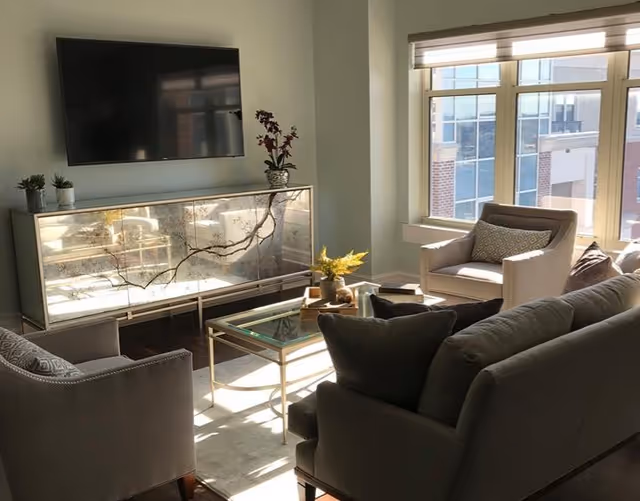 A bright living room with large windows letting in natural light. The room features a wall-mounted flat-screen TV above a mirrored cabinet, a glass coffee table with decorative items, a gray sofa with cushions, and two armchairs with patterned pillows.