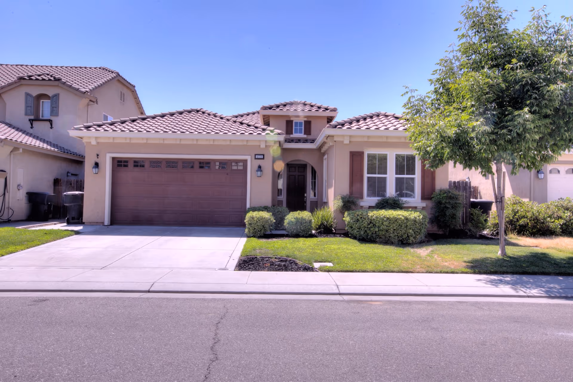 Front exterior view of a single-story house with a tiled roof, a two-car garage with a brown door, a small front porch, and a well-maintained lawn with bushes and a tree.