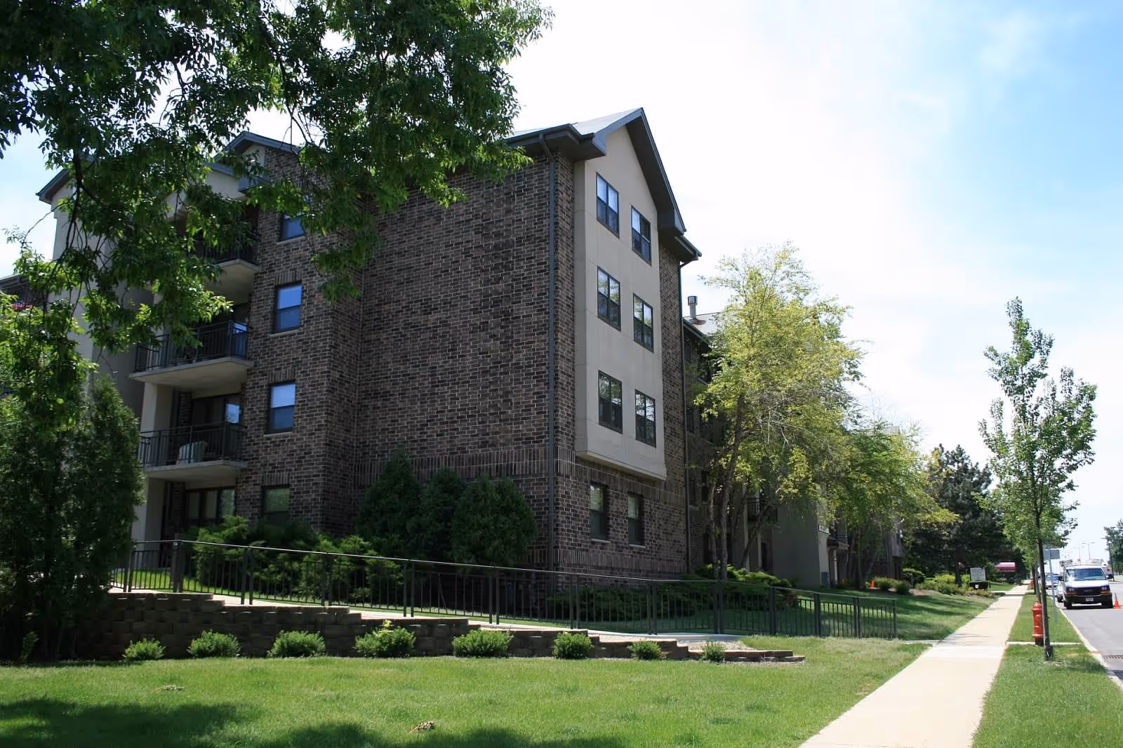Exterior view of a multi-story brick and beige building with balconies, surrounded by green trees and bushes, with a sidewalk and a street running alongside it under a partly cloudy sky.