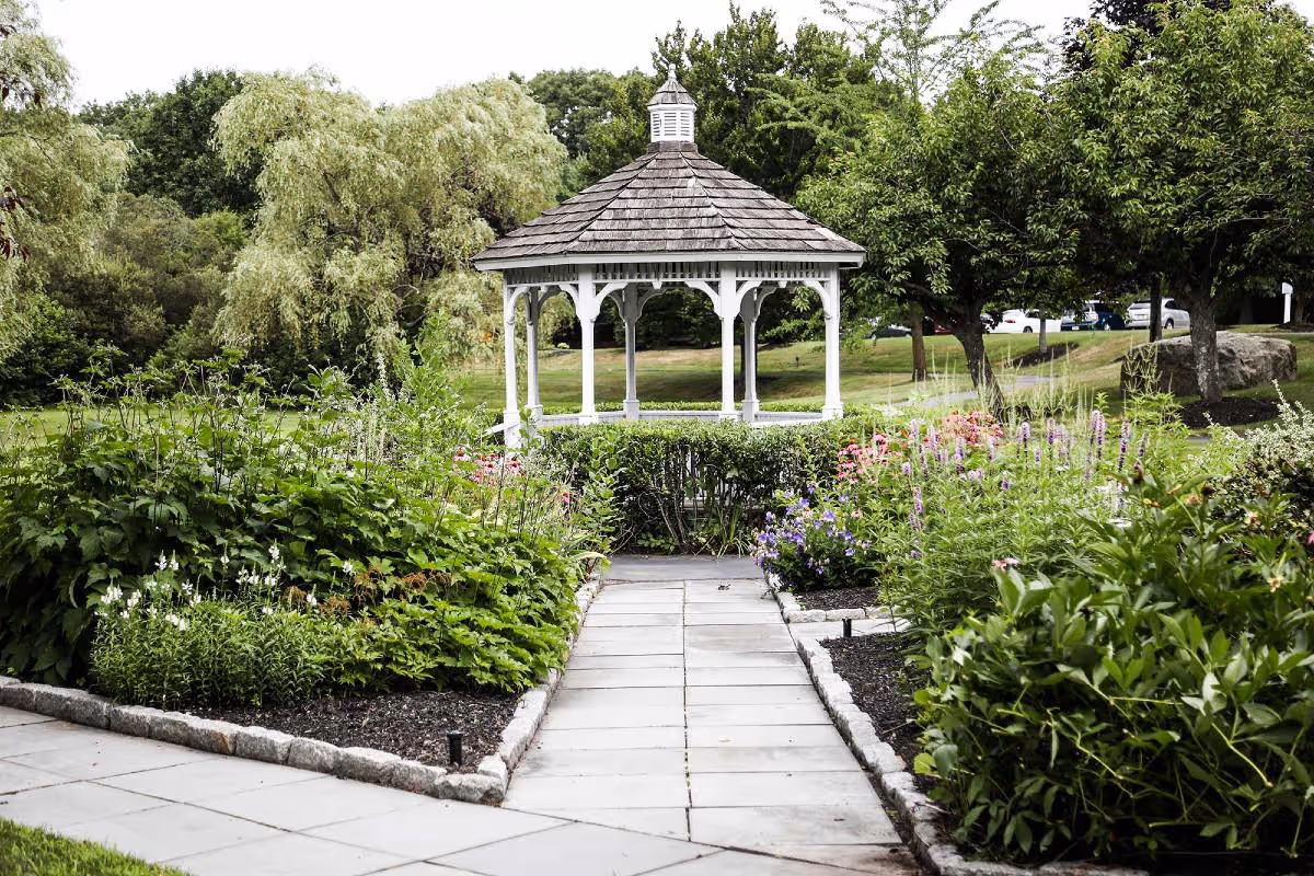 A paved walkway leading to a white wooden gazebo surrounded by lush green bushes, flowering plants, and trees in a garden setting.