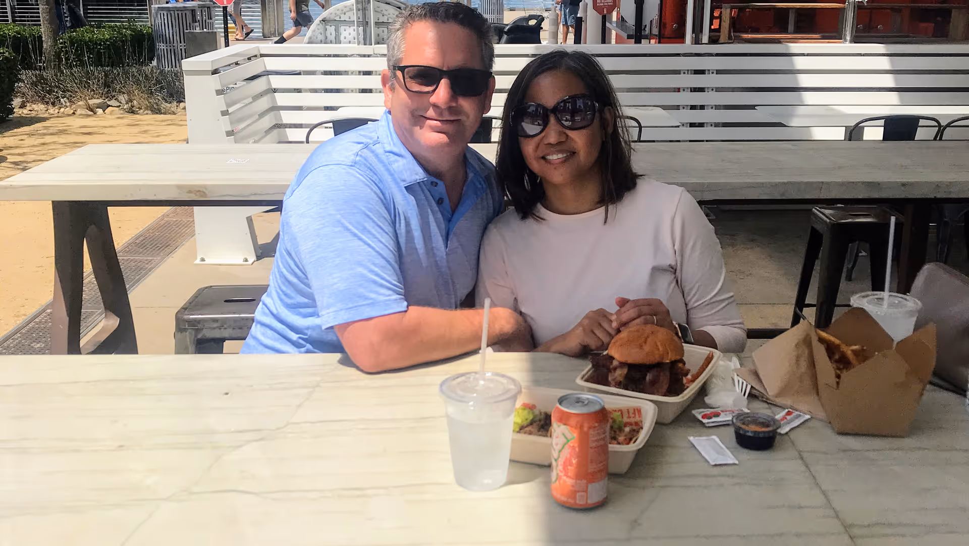 A man and a woman wearing sunglasses sit at an outdoor picnic table with takeout food and drinks.