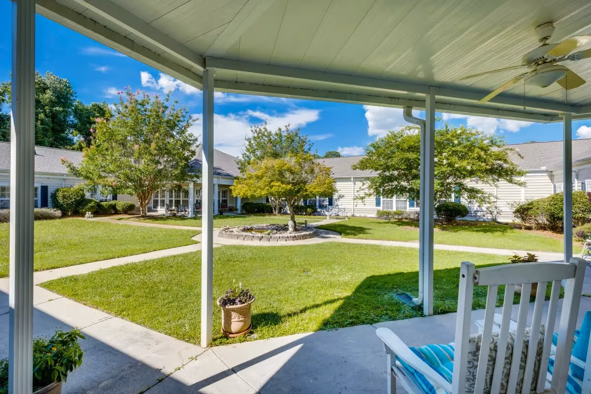 View from a covered patio with a white rocking chair and ceiling fan overlooking a green courtyard with trees, a circular stone planter, and a walking path surrounded by single-story buildings under a blue sky with scattered clouds.