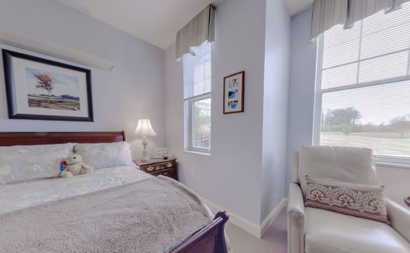 Bright bedroom with a wooden bed topped with a teddy bear, a nightstand and lamp, and an armchair beside two tall windows.
