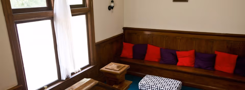 A cozy interior corner with a wooden bench lined with alternating red and purple cushions, a patterned ottoman in front, and a window with sheer white curtains letting in natural light.