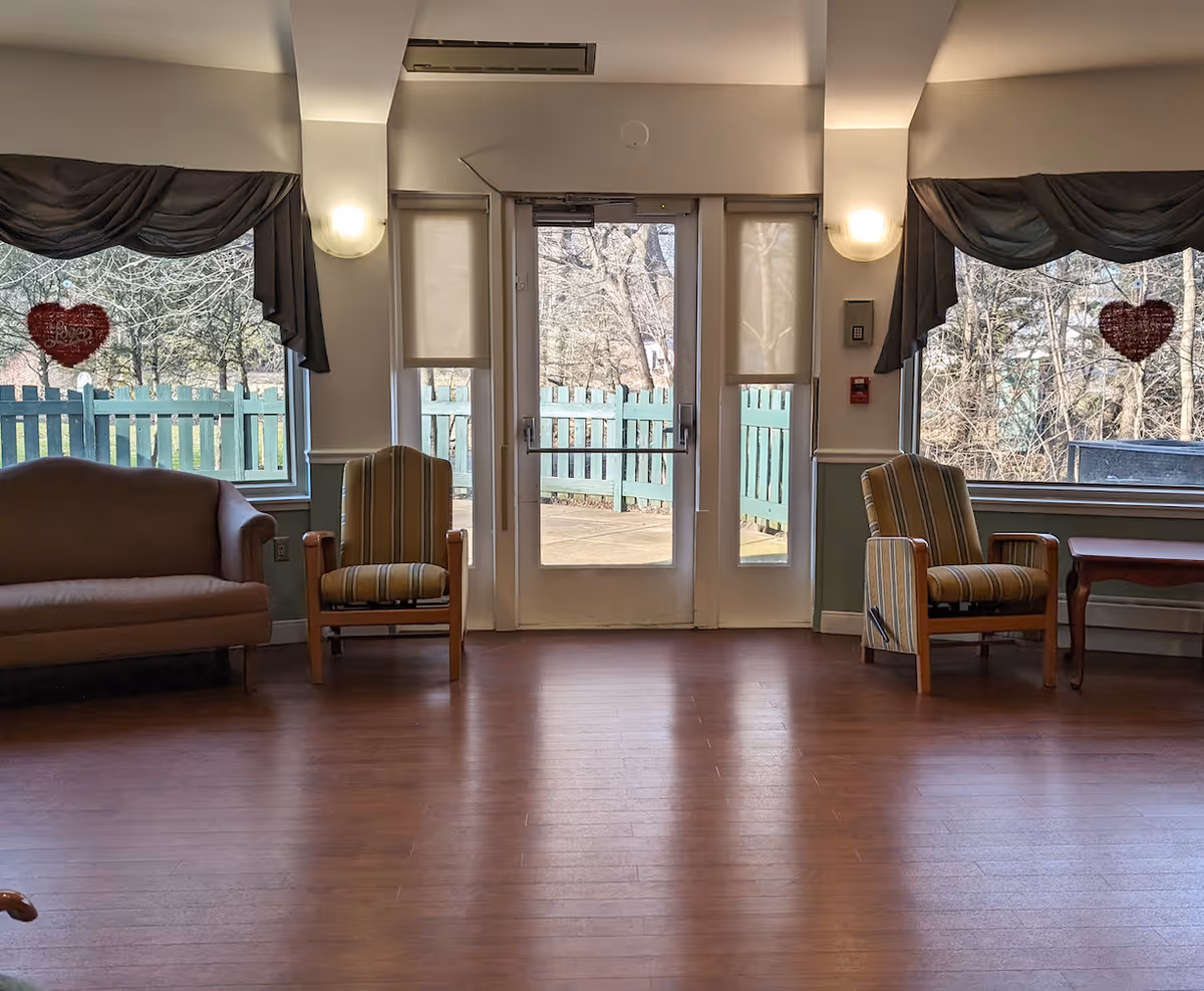 A sitting area inside Naamans Creek Country Manor featuring a brown couch on the left, two striped armchairs, and a wooden table on the right. There are two large windows with dark valances and heart-shaped decorations hanging in each window. A glass door with side windows leads outside to a fenced area with trees visible beyond.