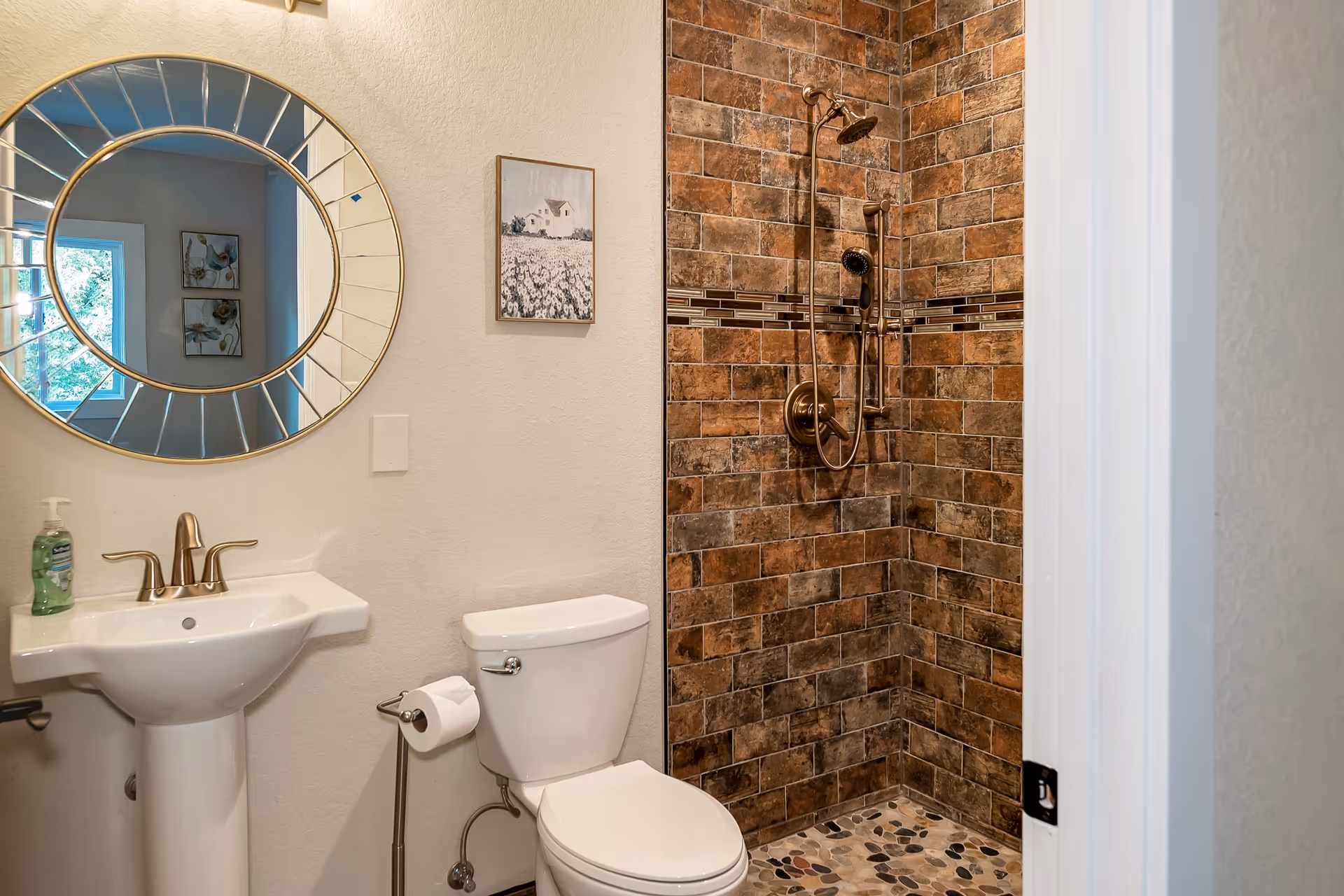 A bathroom with a white pedestal sink, a round decorative mirror above it, a toilet with a toilet paper holder, and a shower area with brown and tan tiled walls and a pebble stone floor. There is a framed picture on the wall between the sink and the shower.
