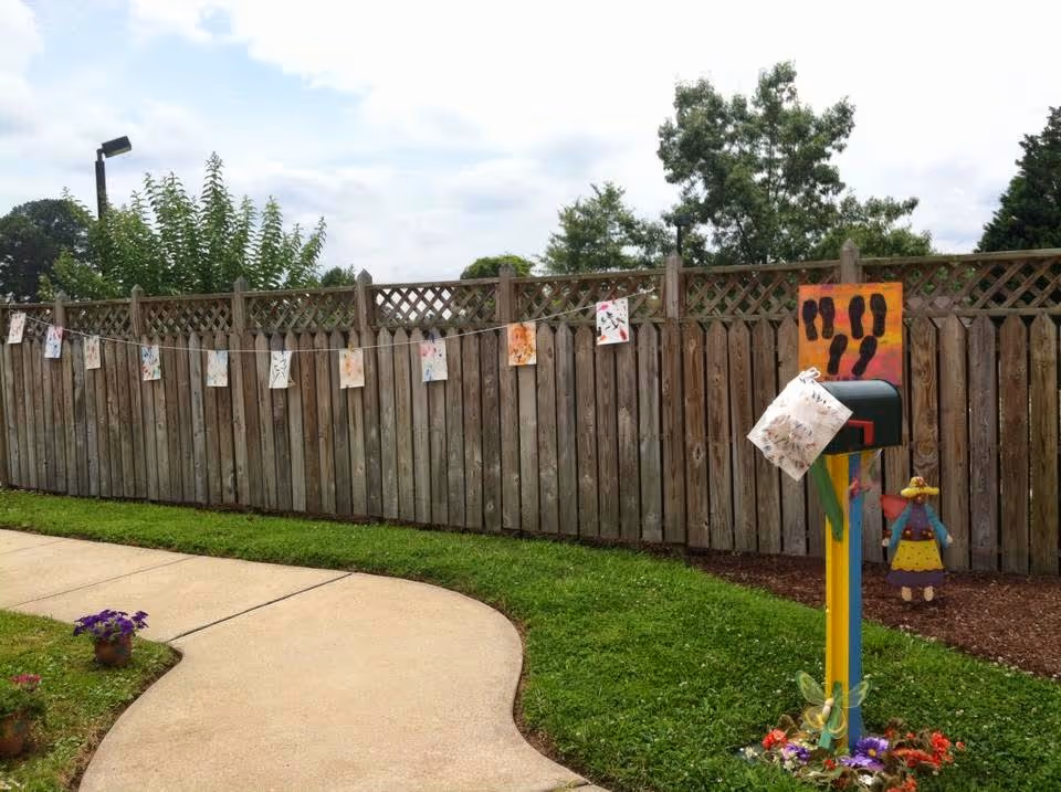 Outdoor area with a curved concrete pathway next to a wooden fence. Various colorful drawings are clipped to a string hanging along the fence. A mailbox painted in bright colors with a butterfly decoration and a small figure is positioned near the pathway. Trees and a cloudy sky are visible in the background.
