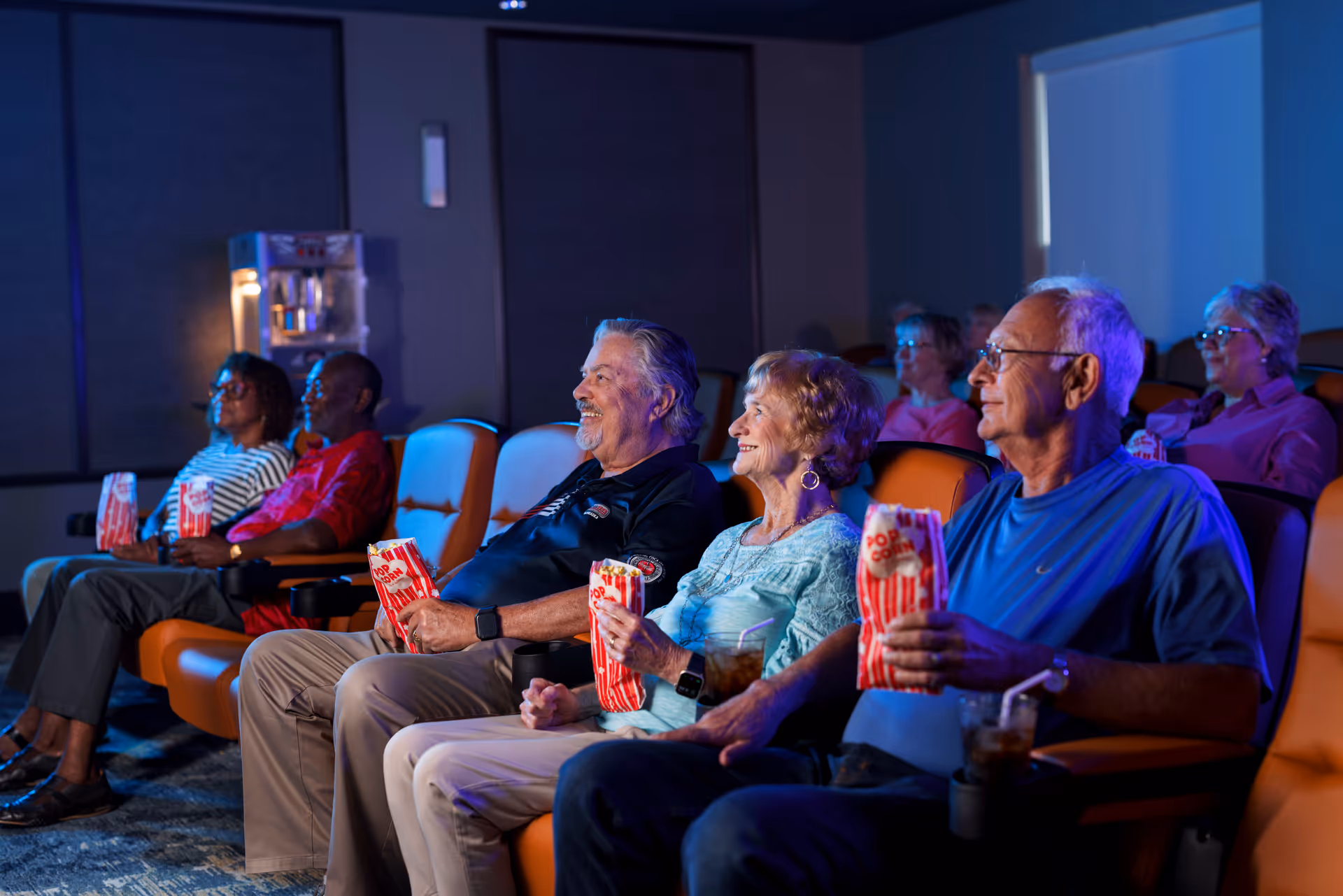 A group of elderly people sitting in a theater-style room watching a movie. They are seated in comfortable orange chairs, holding popcorn and drinks, and appear engaged and happy.