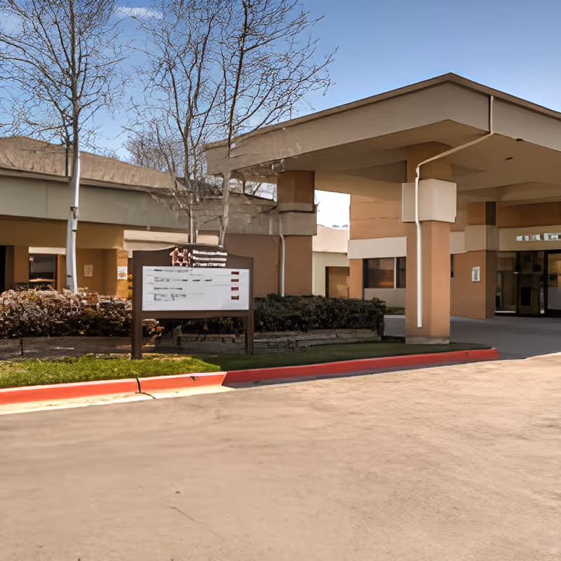 Exterior view of Monument Health Murray Creek facility entrance with a covered drop-off area, a signboard, leafless trees, and some bushes under a clear blue sky.