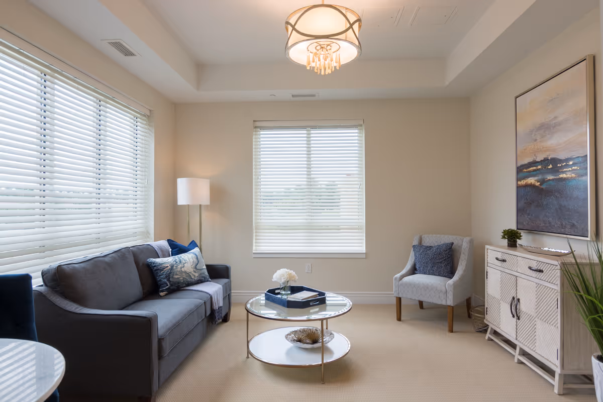 A bright and cozy living room with a gray sofa adorned with blue and patterned pillows, a round glass coffee table with decorative items, a light gray armchair with a blue pillow, a white wooden cabinet with a small plant and decorative tray, a large abstract painting on the wall, and two windows with white blinds letting in natural light.