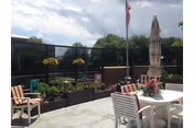 Outdoor patio area with white tables and chairs, some with striped cushions, potted plants and flowers along the fence, an American flag on a pole, and a closed umbrella under a partly cloudy sky.