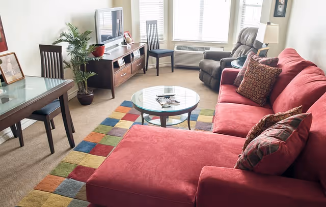 A bright living room with a large red sectional sofa adorned with patterned pillows, a glass coffee table on a colorful checkered rug, a wooden TV stand with a flat-screen TV, a potted plant, a recliner chair, and a dining table with chairs near a window with blinds.