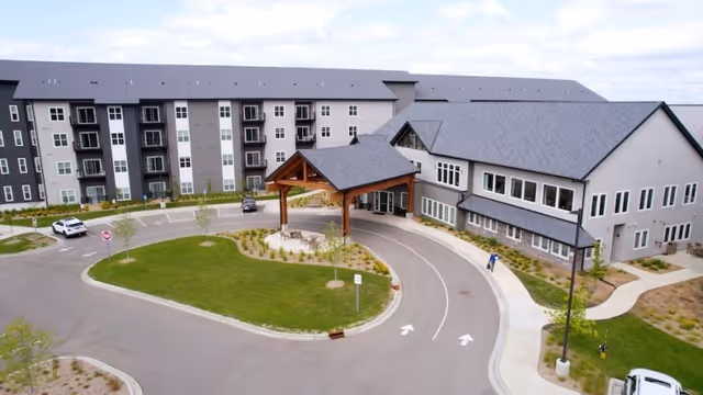 Aerial view of the entrance to Benedictine Living Community- Shakopee Windermere Way, showing a large building with multiple floors, a covered drop-off area, a curved driveway, landscaped green areas, and a few parked cars.