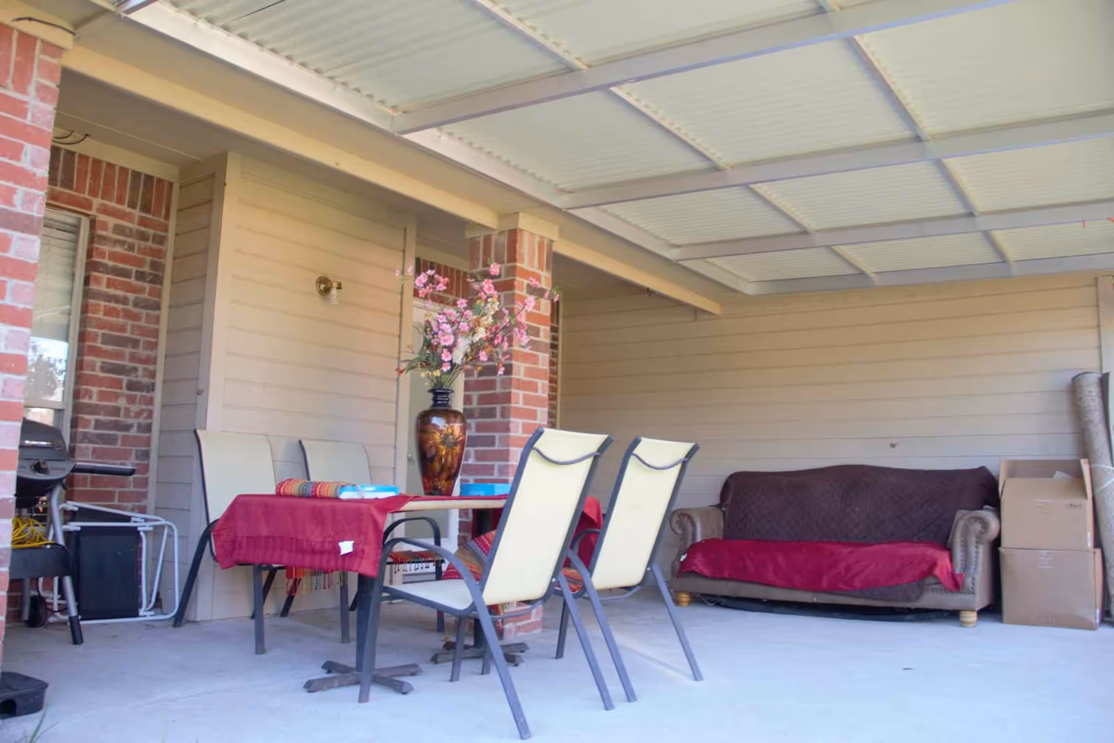 Covered outdoor patio area with a table covered by a red tablecloth, four chairs, a large vase with pink flowers on the table, a brown couch with a red cover, some cardboard boxes, and a barbecue grill in the corner.