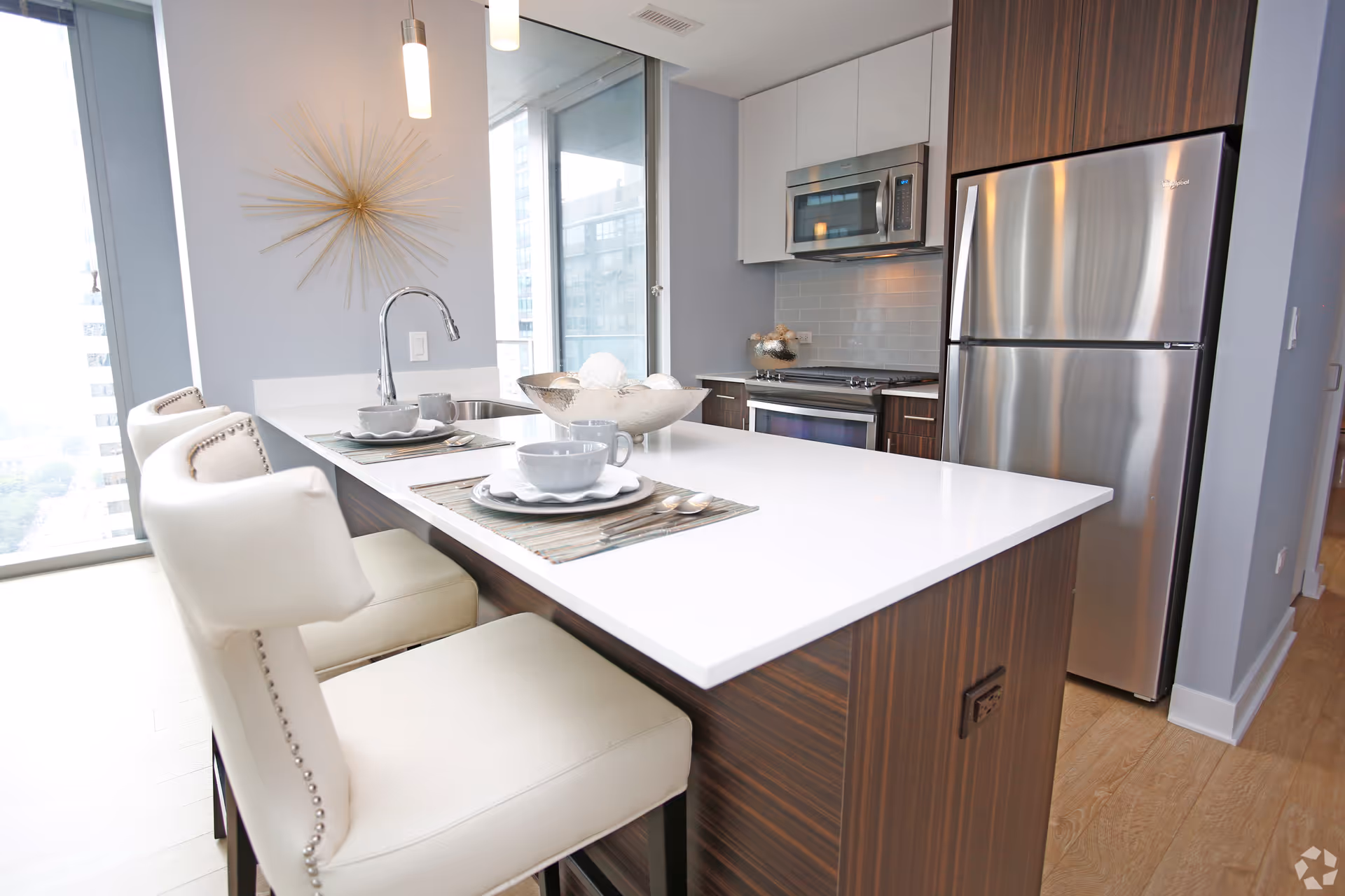 Modern kitchen with a white countertop island featuring two white cushioned bar stools, place settings with bowls, plates, mugs, and silverware. The kitchen has stainless steel appliances including a refrigerator, oven, and microwave. There is a decorative gold wall art piece and large windows providing natural light.